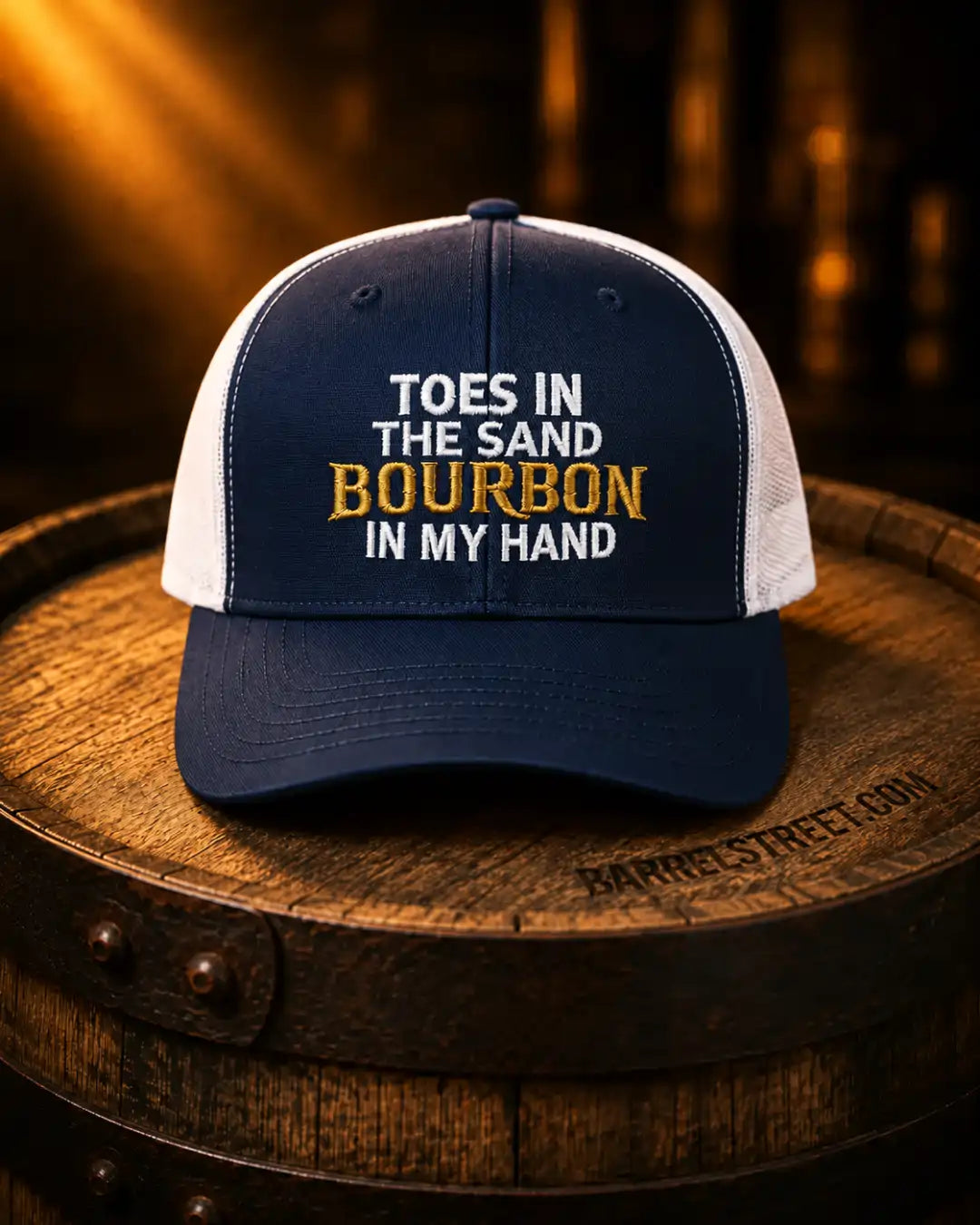 A navy blue and white trucker-style cap with embroidered text reading 'TOES IN THE SAND BOURBON IN MY HAND' sits on a wooden barrel in a warm, dimly lit setting.