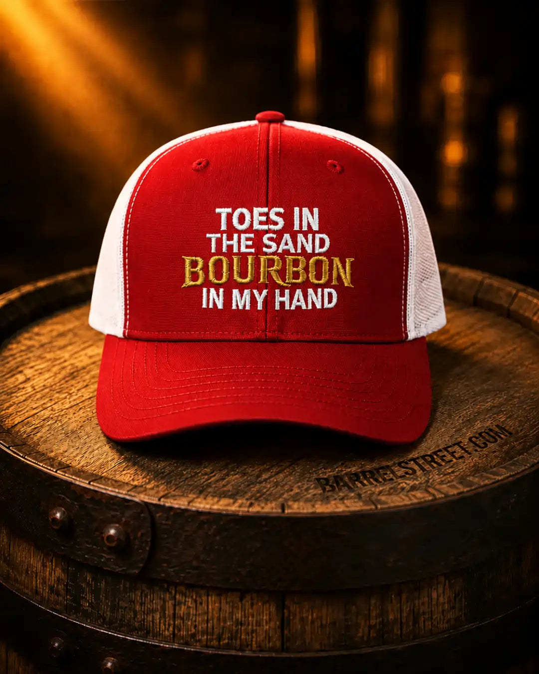 A red and white trucker hat with embroidered text reading 'TOES IN THE SAND BOURBON IN MY HAND' sits on a wooden barrel in a warm, dark setting.