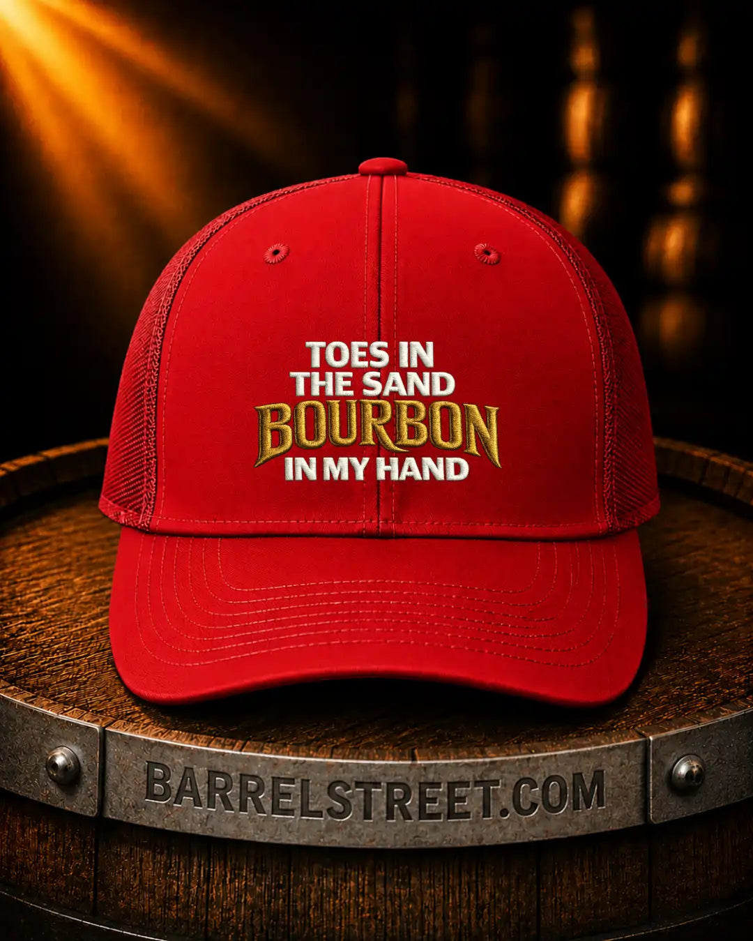 A red trucker-style cap with embroidered text reading 'TOES IN THE SAND BOURBON IN MY HAND' sits on a wooden barrel. The barrel has 'BARRELSTREET.