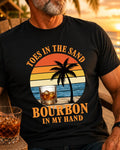 A man wearing a black graphic t-shirt with a retro sunset beach design and the text 'TOES IN THE SAND BOURBON IN MY HAND'.