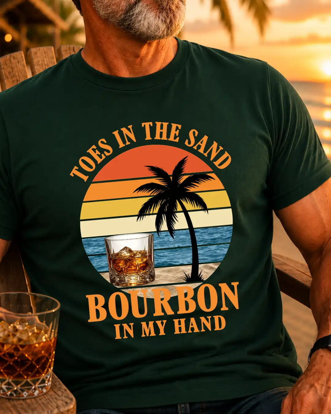 A man wearing a dark green graphic t-shirt with a retro sunset beach design and the text 'TOES IN THE SAND BOURBON IN MY HAND'. A glass of whiskey is visible on a wooden table in the foreground, with a warm sunset beach background.