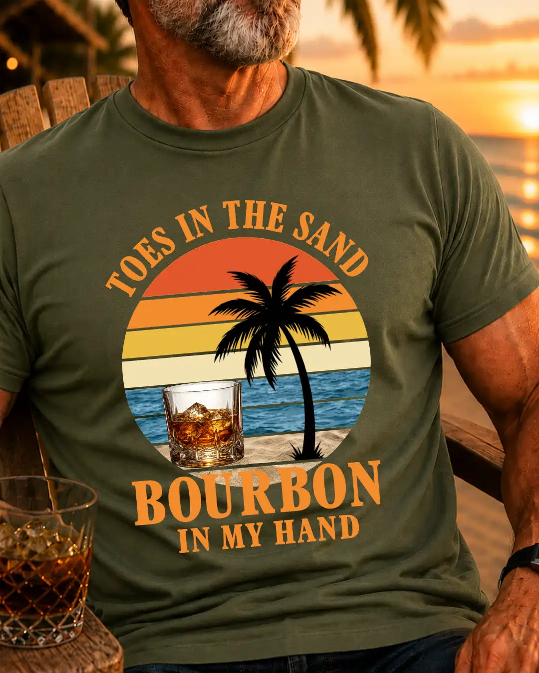 A man wearing an olive green t-shirt with a retro-style graphic that reads 'TOES IN THE SAND BOURBON IN MY HAND' featuring a sunset, palm tree, and a glass of bourbon. The background shows a warm sunset over water.