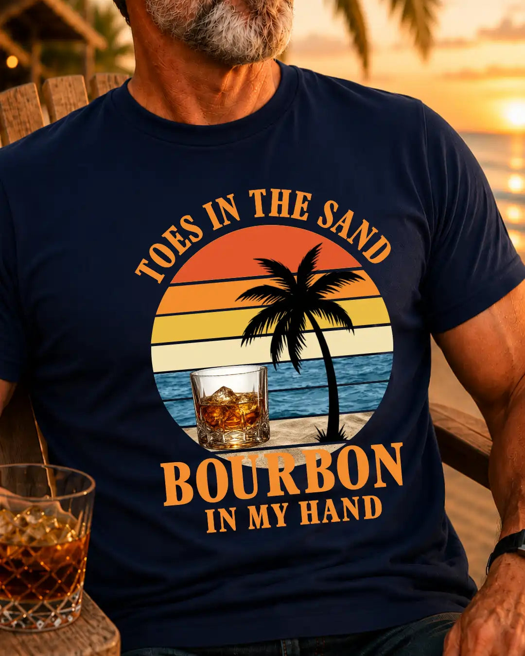 A man wearing a navy blue graphic t-shirt with a retro sunset beach design and the text 'TOES IN THE SAND BOURBON IN MY HAND'. The background shows a warm sunset beach scene.