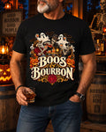 A man wearing a black graphic t-shirt with a Halloween-themed design featuring skeletons, ghosts, pumpkins, and the text 'Boos & Bourbon'. The background is a dimly lit, rustic Halloween-themed room with barrels, lanterns, and skeletons.