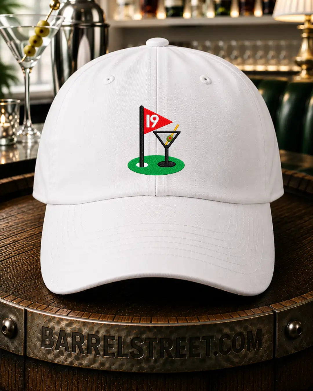 A white baseball cap featuring a golf-themed graphic on the front with a green putting green, a red flag, and a martini glass, displayed on a wooden barrel in a bar setting.