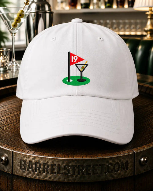A white baseball cap featuring a golf-themed graphic on the front with a green putting green, a red flag, and a martini glass, displayed on a wooden barrel in a bar setting.