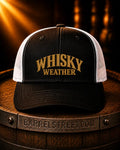A black and white trucker hat with 'WHISKY WEATHER' embroidered in gold on the front, resting on a wooden barrel with a warm, dramatic background.