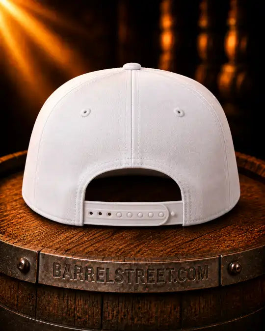 A white structured snapback cap shown from the back, resting on a wooden barrel with a dark, warm-toned background and light rays.